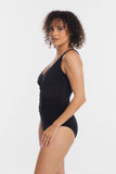 A woman with curly brown hair models a black one-piece swimsuit or bodysuit with a deep V-neckline and gathered ruching on the side, shown from a three-quarters back view against a plain white studio background.
