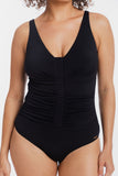 A woman wearing a black one-piece swimsuit with a deep V-neckline, wide shoulder straps, and a vertical gathered seam down the center front, photographed against a plain light background.