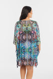 A woman viewed from the back wearing a loose, short-sleeve kaftan-style dress with a colorful abstract and floral print in blue, green, pink, black, and white, set against a plain white studio background.