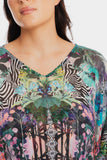 A close-up of a woman wearing a V-neck top with a complex multicolor print featuring tropical leaves, zebra stripes, and floral motifs in green, pink, blue, black, and white.