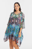A woman modeling a loose, flowy kaftan-style dress with a vibrant multicolor abstract print in shades of blue, green, purple, pink, and black, set against a plain light gray studio background.