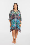 A woman modeling a loose, short-sleeve kaftan-style dress with a vibrant multicolor abstract and floral pattern in shades of blue, green, pink, and black, set against a plain white studio background.
