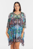 A woman modeling a short, loose V-neck dress with three-quarter sleeves, featuring a vibrant, symmetrical multicolor abstract print in shades of blue, green, pink, purple, and black, set against a plain light gray background.