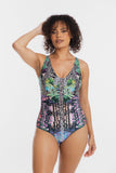 A woman modeling a one-piece swimsuit with a complex multicolor print featuring zebra stripes, green tropical leaves, and pink floral elements on a light gray background.