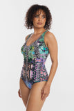 A woman modeling a one-piece swimsuit with a vibrant, multicolor abstract print featuring black, green, pink, purple, and blue tones, set against a plain light gray background.