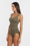 A woman modeling a one-piece swimsuit with a textured, geometric chevron-like pattern in olive green and brown tones, set against a plain light gray studio background.