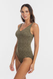 A woman modeling a one-piece swimsuit with a textured chevron pattern in olive green, set against a plain light gray studio background.