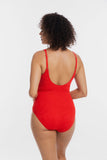 A woman with curly dark hair is shown from the back wearing a bright red one-piece swimsuit with thin shoulder straps and a low open back, photographed against a clean light gray studio background.