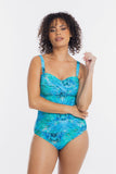 A woman modeling a one-piece swimsuit with a vibrant blue and green tropical leaf pattern against a plain white studio background.