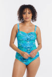A woman modeling a one-piece swimsuit with a vibrant blue and turquoise abstract pattern against a plain white studio background.