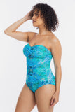 A woman modeling a strapless one-piece swimsuit with a tropical leaf print in shades of turquoise, blue, and teal against a plain white studio background.