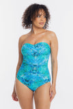 A woman modeling a strapless one-piece swimsuit with a vibrant blue and green tropical leaf pattern against a plain white background.