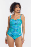 A woman modeling a one-piece swimsuit with a vibrant blue and green abstract pattern, set against a plain light gray background.