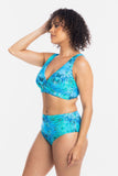 A woman modeling a two-piece bikini swimsuit with a vibrant blue and green tropical leaf pattern against a plain white studio background.
