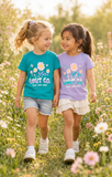 Two young girls walking hand-in-hand through a sunny field of wildflowers, wearing matching graphic t-shirts from Unit Co. One shirt is teal blue with a yellow and white flower design, and the other is lavender purple with a pink and white flower design.