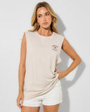 A woman modeling a beige sleeveless crop top with a small dark graphic on the left chest, paired with white textured shorts, against a light gray studio background. Unit Ladies Muscles Tank - Lodge