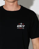 A close-up of a person wearing a black short-sleeve crewneck t-shirt with a white and red graphic on the left chest that reads 'UNIT' with smaller text above and below.