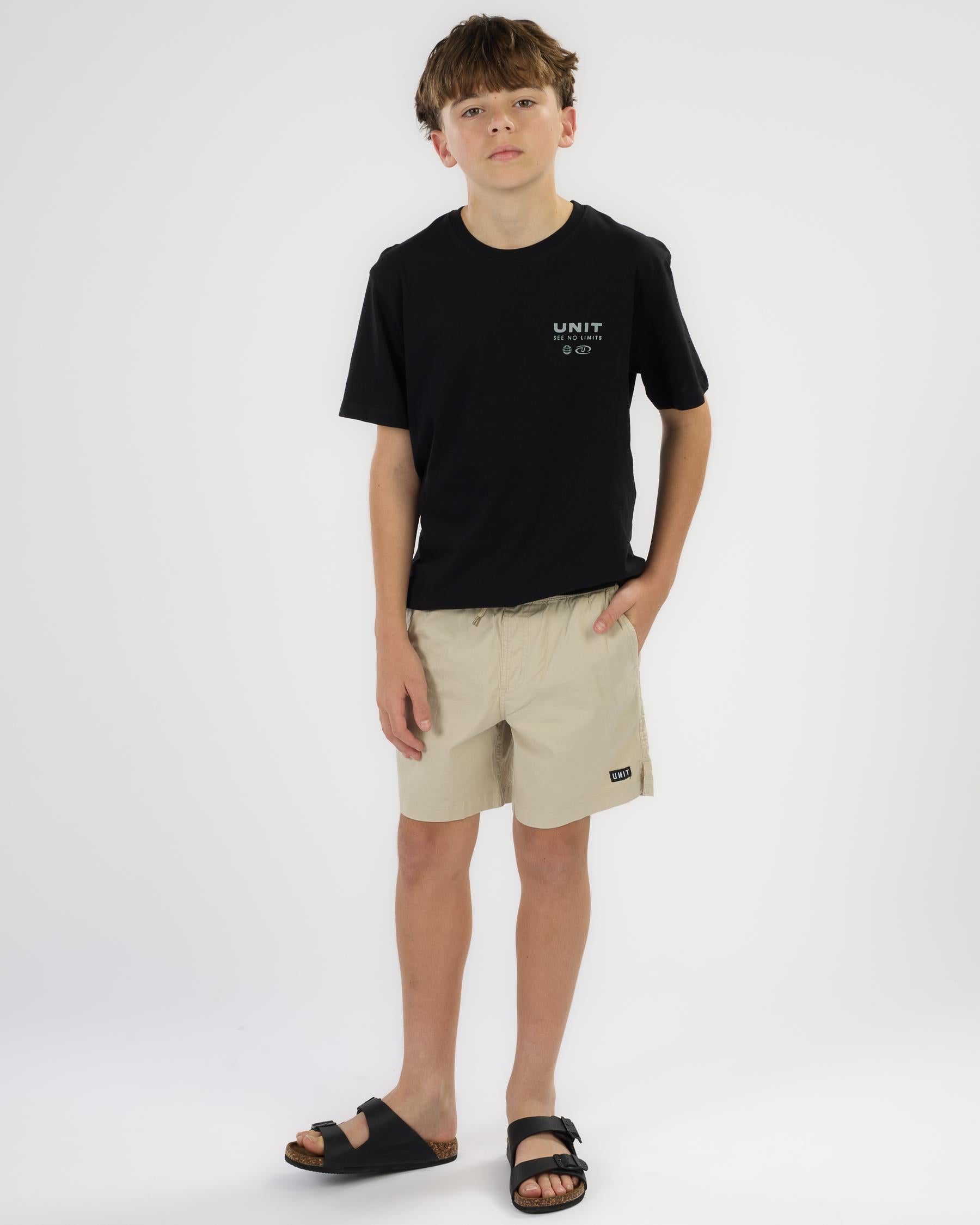 A young boy modeling a black short-sleeve t-shirt with a small white graphic on the left chest, paired with beige shorts and black slide sandals, standing against a plain white studio background.
