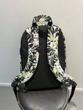 A backpack with a black base and a tropical floral pattern in white, green, and light pink, shown from a side angle against a plain light gray wall.