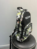 A small shoulder bag with a black base and a tropical floral print in white, green, and soft pink, featuring the brand name 'WILSON' in white on the black section. The bag is displayed on a black leather saddle against a plain light gray background.