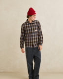 A model wearing a brown and black plaid button-down shirt with a small Carhartt logo patch on the chest, paired with dark gray pants and a red beanie, standing against a textured beige studio backdrop. Rhythm Mens Alex Check LS Shirt - Chestnut