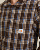 Close-up of a brown, beige, and blue plaid button-up shirt with a small orange and white Carhartt logo patch on the chest pocket. Rhythm Mens Alex Check LS Shirt - Chestnut
