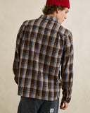 A man wearing a brown, beige, and blue plaid flannel shirt, dark jeans, and a red beanie, shown from a back-three-quarter view against a neutral beige background. Rhythm Mens Alex Check LS Shirt - Chestnut