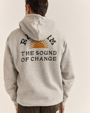 A man viewed from behind wearing a light heather gray hoodie with a large graphic on the back. The graphic features a yellow-orange rising sun and the text 'RHYTHM THE SOUND OF CHANGE' in black. Rhythm Mens Awake Fleece Hood - Grey Heather