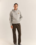 A man wearing a light gray pullover hoodie with a small orange and yellow graphic on the left chest, paired with dark brown pants and black-and-white sneakers, standing against a plain light gray studio background.
