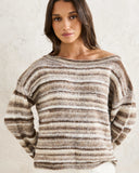 A woman wearing a fuzzy, off-the-shoulder knit sweater with horizontal stripes in brown, beige, and cream, standing against a textured off-white wall. Front view of the neutral sand Rhythm Baklava Knit sweater showing the relaxed fit, crew neck, and textured open crochet weave.