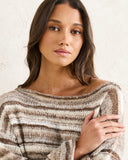 A woman wearing a chunky knit off-the-shoulder sweater with horizontal stripes in brown, beige, and cream, posing against a textured off-white wall. Rhythm Baklava Knit - Sand