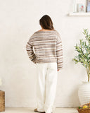A woman viewed from behind wearing a brown and beige striped knit sweater and white wide-leg pants, standing in a bright room with a textured white wall, a potted plant, and a small shelf. Rhythm Baklava Knit - Sand