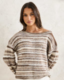 A woman wearing a fuzzy, off-the-shoulder knit sweater with horizontal stripes in brown, beige, and cream, standing against a textured off-white wall. Rhythm Baklava Knit - Sand