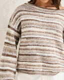 A close-up of a person wearing a chunky knit sweater with horizontal stripes in brown, beige, and cream, paired with white pants against a textured light wall. Rhythm Baklava Knit - Sand