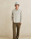A man wearing a light beige long-sleeve button-up shirt, olive brown trousers, and a light blue cap with a green logo, standing against a textured beige studio wall. Rhythm Men's Bank Stripe LS Shirt – Olive