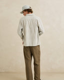 A man wearing a light beige linen button-up shirt, brown trousers, and a light blue cap, photographed from behind against a textured beige wall. Rhythm Men's Bank Stripe LS Shirt – Olive