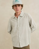 A man wearing a light beige and white vertically striped button-down shirt, a white cap with green text, and brown pants, standing against a neutral beige background. Rhythm Men's Bank Stripe LS Shirt – Olive