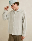 A man wearing a light beige and white vertically striped long-sleeve button-up shirt, brown pants, and a light gray baseball cap, posing against a neutral beige textured wall. Rhythm Men's Bank Stripe LS Shirt – Olive