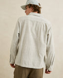 A man wearing a light beige and white vertically striped long-sleeve button-up shirt, brown pants, and a light blue cap, shown from a back-three-quarter view against a neutral beige background. Rhythm Men's Bank Stripe LS Shirt – Olive