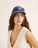 Front view of the Rhythm Bayside Cap in Washed Dark Blue showing the low-profile six-panel crown, curved brim, and cream embroidered front logo. A woman wearing a blue denim baseball cap with a white embroidered logo patch, paired with a light beige sleeveless top, set against a plain beige background.