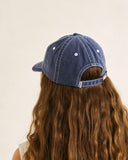 A person shown from the back wearing a blue denim baseball cap with a curved brim and adjustable strap, against a plain light beige background. Back view of the Rhythm Bayside Cap in Washed Dark Blue highlighting the matching fabric adjustable strap and metal slide buckle setup.