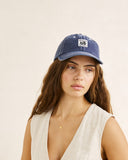 A woman wearing a blue denim baseball cap with a white embroidered logo patch, paired with a light beige sleeveless top, set against a plain beige background.
