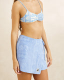 A woman wearing a light blue patterned bikini top and a matching light blue textured wrap skirt with a small white logo patch near the hem, photographed against a plain beige studio background. Rhythm Bayside Terry Towelling Wrap Mini Skirt