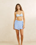 A woman modeling a light blue patterned bikini top and matching light blue swim shorts against a warm beige studio backdrop. Rhythm Bayside Terry Towelling Wrap Mini Skirt