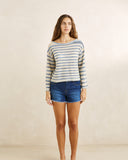 A woman modeling a long-sleeve knit top with thin blue and beige horizontal stripes, paired with blue denim shorts, standing against a neutral beige textured wall. Rhythm