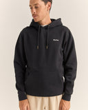 A man wearing a black pullover hoodie with a small white 'Rhythm.' logo on the left chest, paired with light beige pants, photographed against a plain light beige studio background. Rhythm Mens Brand Fleece Hood - Black