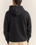 A person shown from the back wearing a plain black pullover hoodie against a light beige studio background. Rhythm Mens Brand Fleece Hood - Black
