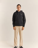 A man modeling a black pullover hoodie with a small white embroidered logo on the left chest, paired with beige trousers and black shoes, against a plain light beige studio background. Rhythm Mens Brand Fleece Hood - Black