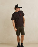 A man wearing a dark brown t-shirt with a small red and yellow graphic on the left chest, paired with olive green shorts, a black and white trucker hat, and black socks, standing against a textured beige wall. Rhythm Mens Cactus SS T-Shirt - Vintage Black