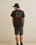 A man wearing a dark brown t-shirt with a colorful desert landscape graphic on the back, paired with olive green shorts and a black baseball cap, standing against a textured beige wall. Rhythm Mens Cactus SS T-Shirt - Vintage Black